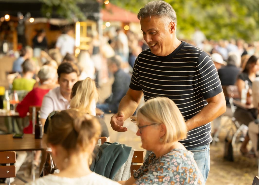 Gastgeber im Ringelshirt serviert Gästen an der langen Tafel im Freien